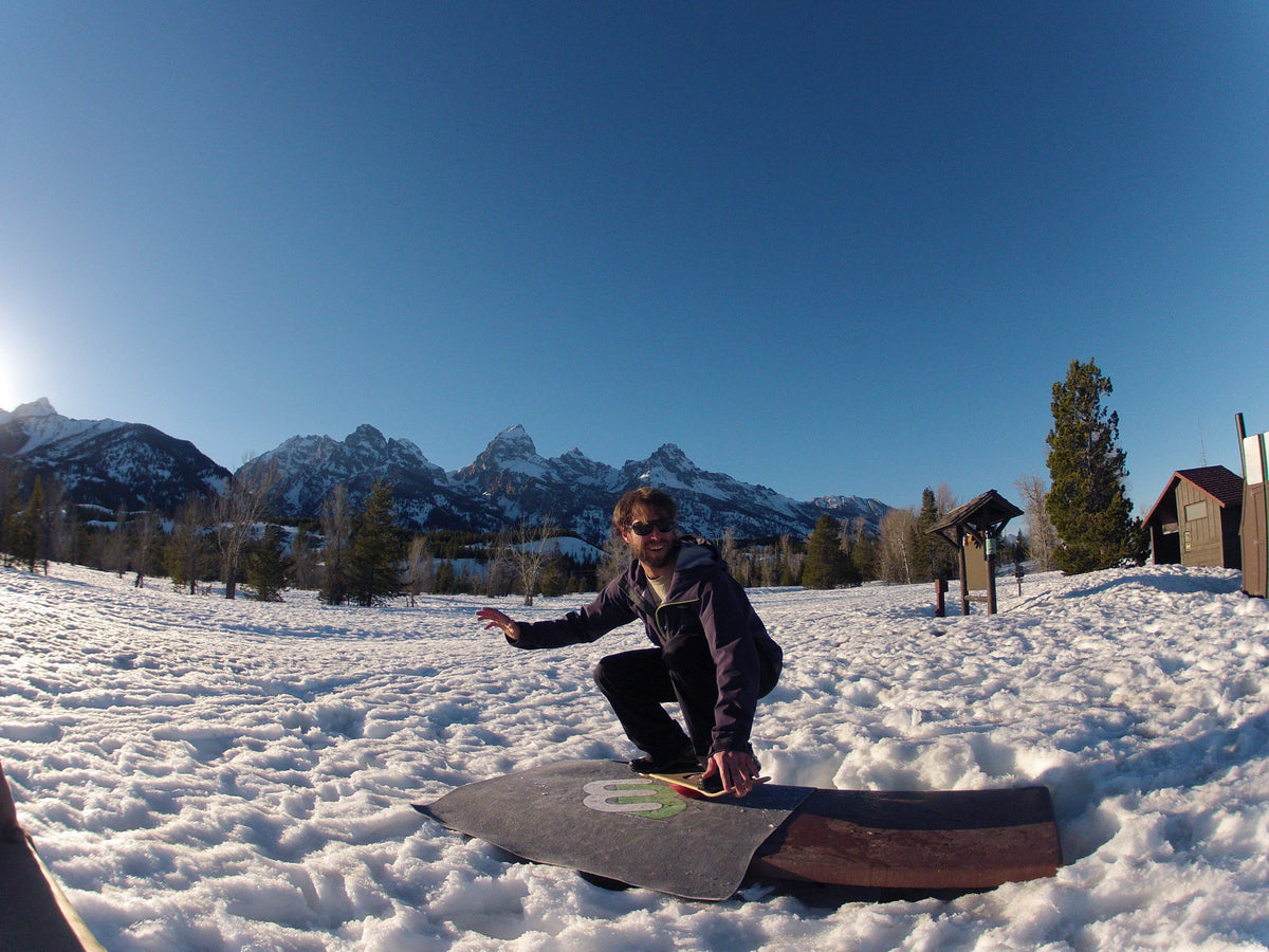Man kneeling on a Whirly Board in the snow with mountains behind