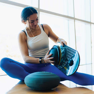 Woman switching balance board attachments showing progressive trainer with ball and cushion accessories